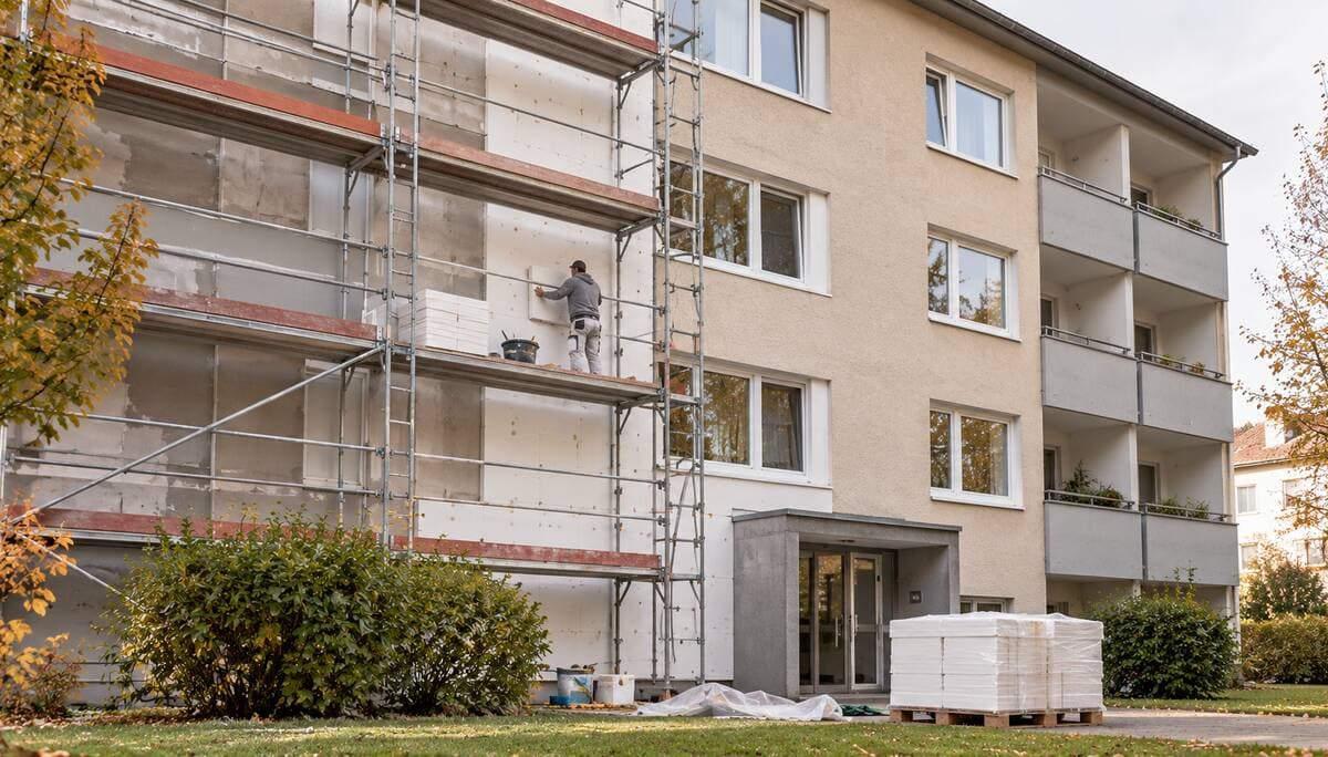 Mieterhöhung nach Modernisierung: Wohnblock mitten in der energetischen Modernisierung, linke Hälfte eingerüstet, ein Handwerker setzt Dämmplatten, davor eine Palette mit Dämmstoff